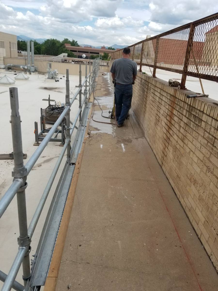 A saw operator walking behind a concrete saw on a rooftop walkway, with a water hose feeding the saw and wet slurry on the concrete